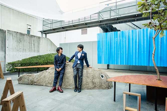 中庭で、魚に見立てた岩に腰掛けるトラフ建築設計事務所の二人。右が鈴野浩一、左が禿真哉。背景の青いカーテンは工事現場で使う養生シートにプリーツ加工したもの。テキスタイルデザイナーの安東陽子とコラボレーションした。