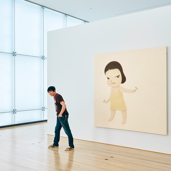 自然光の入る気持ちのいい空間に絵が伸び伸びと展示された部屋で、絵に描かれた少女と似たポーズをとる奈良さん。《Sprout the Ambassador》（2001）個人蔵。
　