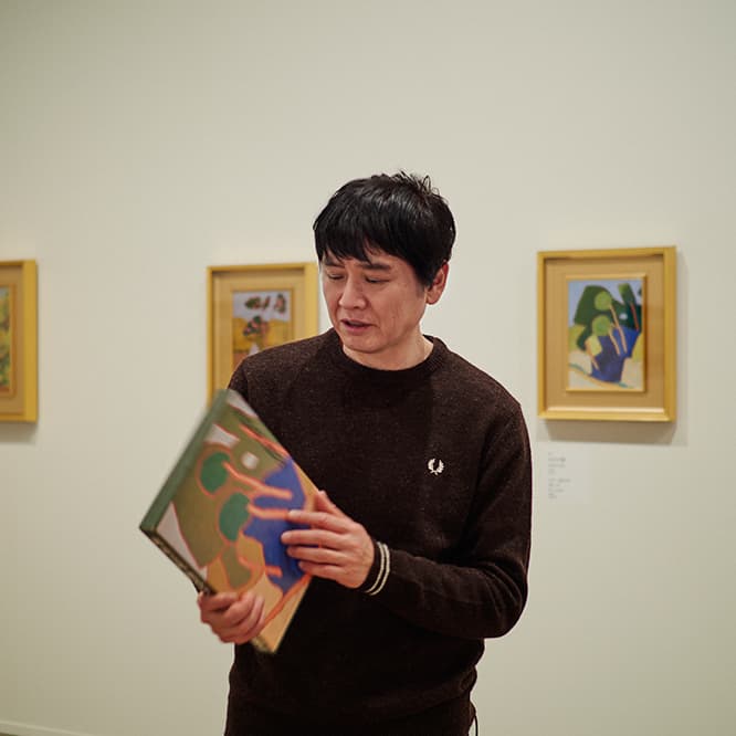 高校生の時にプレゼントされた画集『〈愛蔵普及版〉現代日本美術全集18萬 鉄五郎／熊谷守一』（集英社 1974年）を手に取る奈良美智さん。背景の絵、右側は本の函に使われた《わさび谷》（1958年）の類似作品である《わさび畑》1949年 油彩・板 33.4×24.3cm。