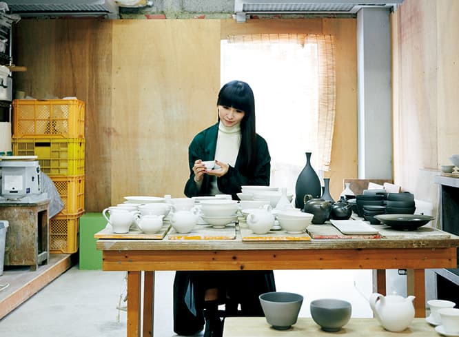 金沢で白磁の作品をつくる森岡希世子さんの工房で。ずらりと並んだボウルやカップを目の前に、真剣な表情で買い付け商品をセレクト中の「かしゆか商店」店主。茶杯には実際にお茶を入れていただき、口あたりを確認。コート98,000円、スカート44,000円（共にY’s／ワイズ プレスルーム TEL03 5463 1540）。トップス、ピアス、靴、靴下はスタイリスト私物。
