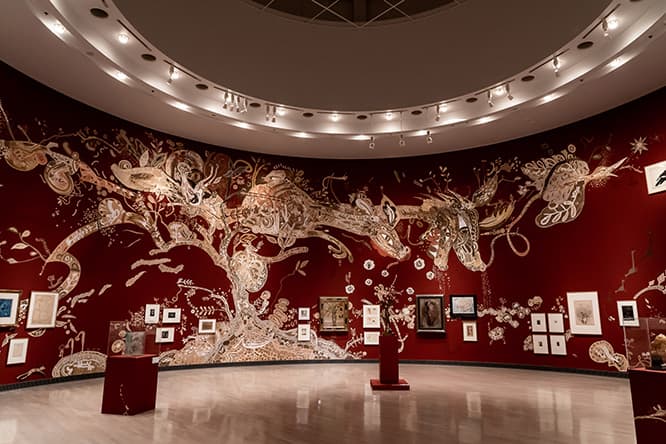 ゲスト・アーティスト、淺井裕介の展示室。円形の展示室の壁いっぱいに淺井が描いた泥絵《いのちの木》（2019年、展示風景）と、彼がセレクトした絵が踊るように並ぶ。