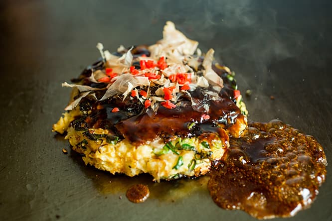「ながおかのお好み焼」1,000円。豚バラはカリカリに焼き上げ、食感のアクセントにもなるコンニャクとジャガイモはあらかじめ煮てある。鉄板で焼く料理は冷めないよう、各席の鉄板の上に運ばれる。