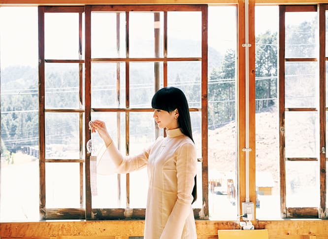 「光が入る場所で見てもいいですか？」と窓際で風鈴を眺めるかしゆか店主。「透明で何も模様のない風鈴は初めて。ガラスの美しさが際立ちますね」。地元の木材や古い建物のガラス窓を再利用した工房の建物にも興味津々。