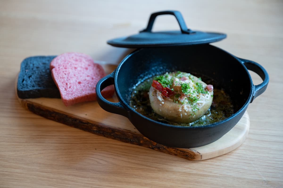 「特大マッシュルームのアヒージョ 竹炭のブルマン添え」（7,500円のプリフィクスメニューの前菜）。肉厚のマッシュルームがオリーブオイルの中でグツグツと煮え立ってくる。竹炭などを練り込んだパンにつけて、召し上がれ。