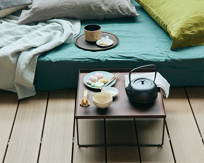 脚付きのお膳やお盆を使えば、好きな場所に茶道具や菓子を持ち運んで楽しめる。内田鋼一の鉄錆膳（私物）の上・重ねた豆皿は各829円、その上のヒメフォークは5本組で4,500円、青い皿の上の菓子切は鈴木務の作。各4,000円、鉄瓶15,000円、ナプキン各 2,000円、マットレスの上の抹茶碗12,000円（以上リビング・モティーフ TEL 03 3587 2784）