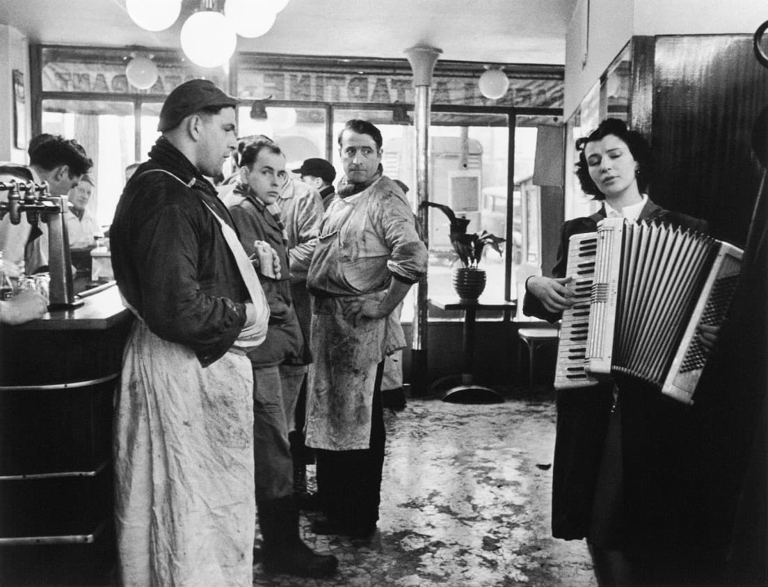 ロベール・ドアノー《音楽好きの肉屋》パリ 1953年2月 ©Atelier Robert Doisneau/Contact。カフェの中、エプロン姿で休憩中の肉屋が、ビエレット・ドリオンの演奏にじっと聞き惚れている。