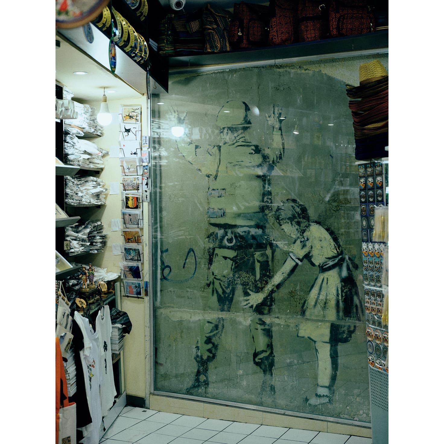 Girl Frisking Soldier(2005)　土産物屋の店内にある作品。バンクシー作品を見学する旅行者が後を絶たないことから、壁を取り込む形で土産物屋がオープン。作品を見学するためには土産物屋に入店する必要があり、入ると商品を勧められる。●Hebron Rd沿いの〈BANKSY SHOP〉という土産物屋内。

