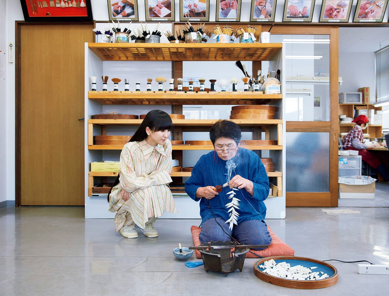 広島の熊野町で熊野筆を制作する〈仿古堂〉。「真っ白でツヤツヤ！」とかしゆか店主も驚いた山羊の毛で、書筆を作る香川翠皐さんと。筆の穂首の根元を焼きごての熱で固めながら、歯で麻糸を引っ張ってしっかりと締める。