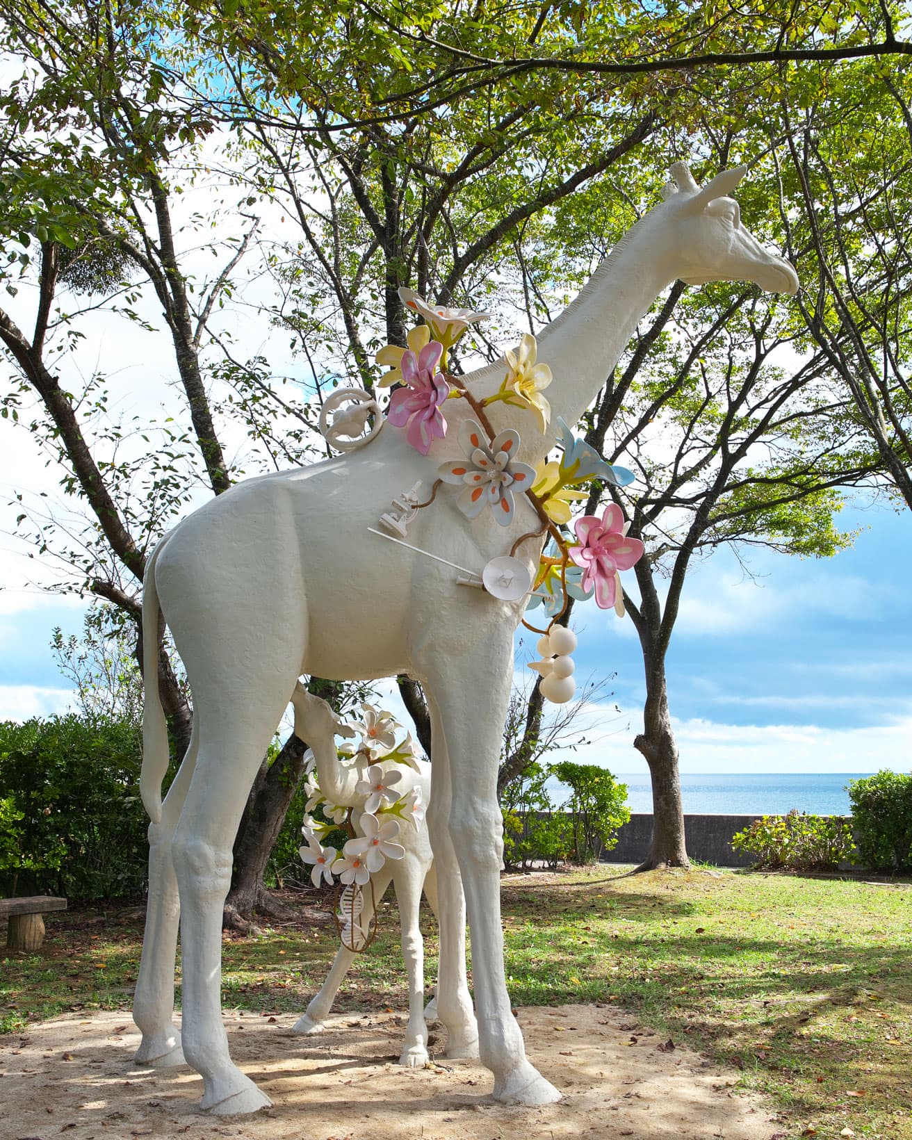 N.S.ハーシャ《なぜここにいるのだろう》。色とりどりの花輪を首にかけて、海を見つめるキリン。　撮影：岡村喜知郎