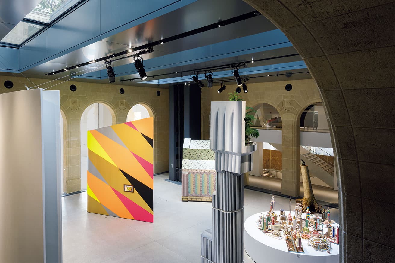 「建築という装置」と題された、第1展示室を、2階のバルコニーから。左端の筒状に見える白い壁は、石上純也の《谷の教会》（2016年）の1/10模型。右端の円形テーブルの上は、コンゴのボディス・キンジュレスによる、ユートピア都市の模型《Projet pour le Kinshasa du troisième millénaire》（1997年）。