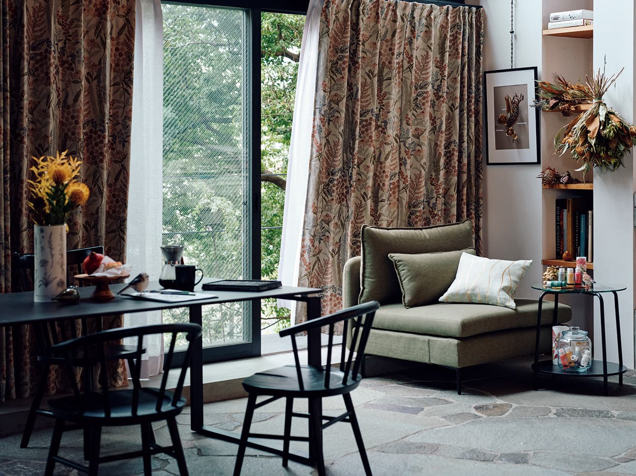 ジャカードカーテンの立体感のある花柄ならば、カーテンがまるで風に揺れる花束となってインテリアの主役になるはず。カーテンとの暮らしがより豊かな時間に。オーダーカーテン1.5倍ヒダ（Ｗ176×Ｈ213㎝×２枚）30,900円、オーダーレースカーテン２倍ヒダ（Ｗ176×Ｈ212㎝×２枚）44,000円、ポスターフレームA2 1,990円、１人用ソファ（Ｗ88×Ｄ98×Ｈ84㎝）44,990円、オーダークッションカバー1,490円、サイドテーブル（Ｗ54×Ｄ54×Ｈ54㎝）8,990円、ガラスジャー999円、ダイニングチェア（手前）各25,000円、ダイニングチェア（奥）14,990円、ダイニングテーブル（Ｗ150×Ｄ90×Ｈ70㎝）79,990円、マグ1,290円、ドリッパーセット2,490円（以上ニトリ）。テーブル、ガラスジャーなどの絵画用品、置物、棚の書籍、飾られた植物などはスタイリスト私物。