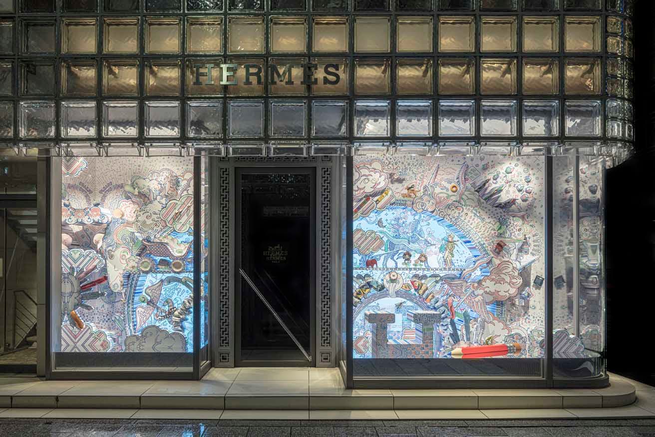 〈銀座メゾンエルメス〉の入口。 レンゾ・ピアノが設計したガラスブロックが印象的な建築は、夜に見ると光を放つ美しいランタンの灯りのよう。このウィンドウの絵は手で描かれている。© Photo by Satoshi Asakawa Created for Maison Hermès Ginza, May 2025
