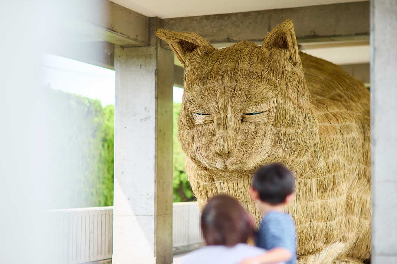 松本勇⾺《スカイネッコ》2025年。〈富山港展望台〉（富山市東岩瀬町海岸通り5）に現れた、藁でできた巨大なネコ。木や竹でできた内部の構造体を作るところから、地域の人とともに作業して制作している。近づいてみると、薄目を開けた目の部分から瞳が見える。松本は同様に藁でできた巨大な牛を〈旧林医院〉（富山市東岩瀬町273）前に展示している。
