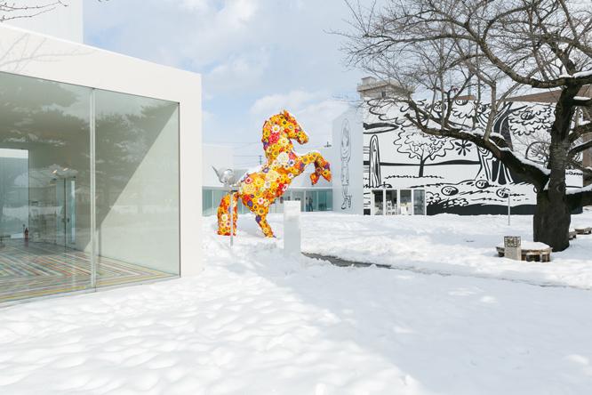 メインエントランス脇に立つ韓国人アーティスト、チェ・ジョンファの《フラワー・ホース》は美術館の顔ともいえる作品。奥の建物の外壁画はポール・モリソン（イギリス）の《オクリア》と奈良美智による《夜露死苦ガール2012》。設計は西沢立衛。Courtesy of Alison Jacques Gallery, London