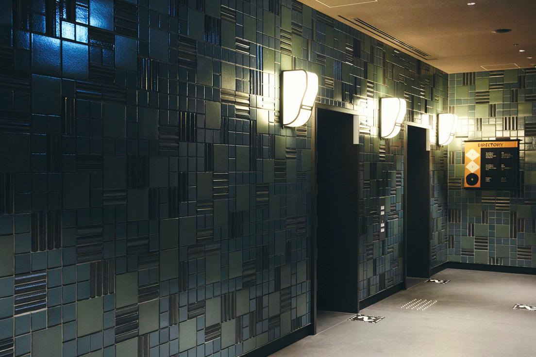 1階エレベーターホールの壁に使用されているのは信楽焼のタイル。サイズや形状の違う複数の種類を組み合わせたブルーやグリーン系の配色は、どことなく藍染の古布にも似た雰囲気。照明の光がタイルに反射して濃淡を強調する。