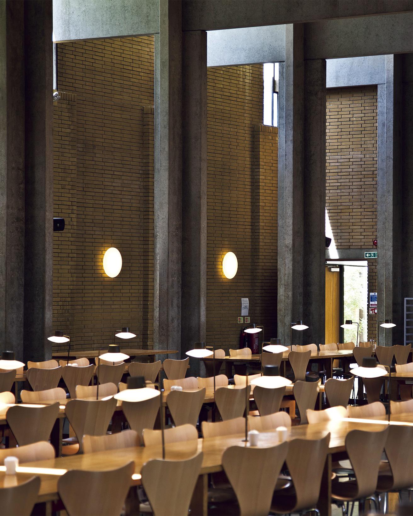コンクリートの十字形の柱と梁が可能にした大空間。オックスフォード大学の中でも一番サイズの大きい350人収容のダイニングホールである。サイドとトップから取り込む自然光を補うのが梁と同じくグリッド状に並ぶ照明だ。レンガの壁にはロードヴル市庁舎用にデザインされた《AJ エクリプタ》が並ぶ。