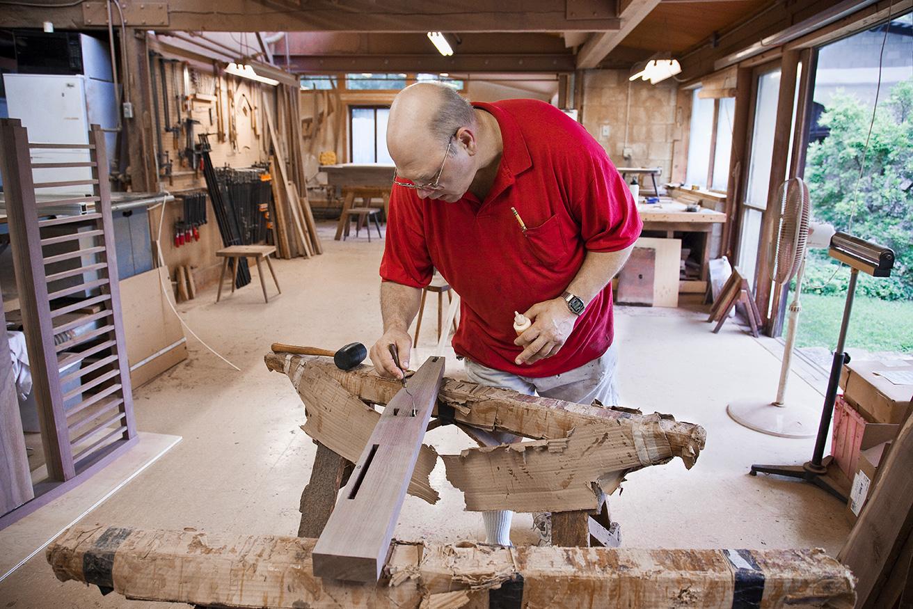 最古参の木匠にして工房の総監督ジェリーが、枠の内側に接着剤を塗る。この後、左手に見える羽根板製ヘッドボードにはめていく。