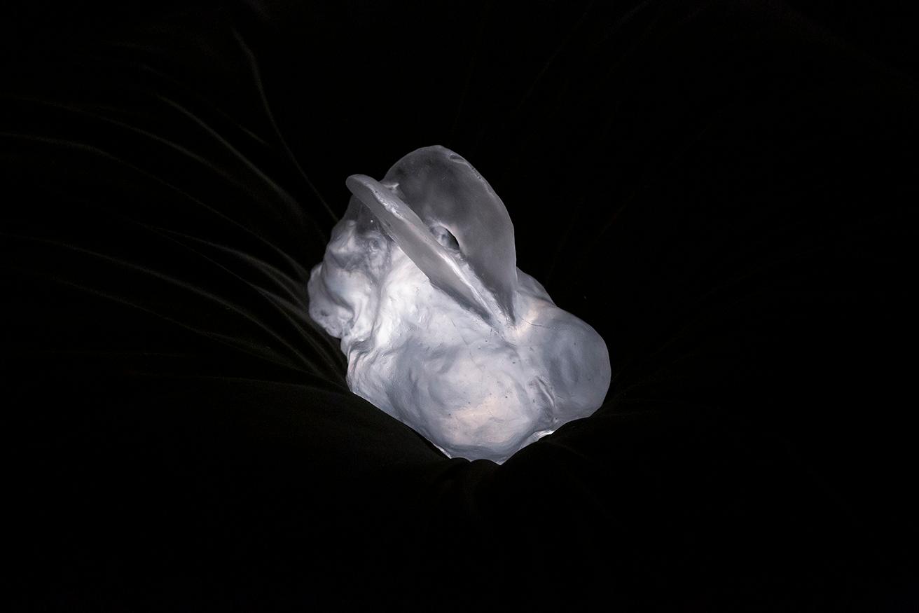 イケムラの作品、透明のウサギはイタリア・ムラノのガラス工房での製作。妥協のないライティングにより、クッションの上にずっしりと存在感をアピールしている。Leiko Ikemura, Transparent Hare, 2022, cast glass (1 / 5), 31×16×17 cm photo_Wai Kung.Courtesy the artist. (c) Leiko Ikemura and The Feuerle Collection